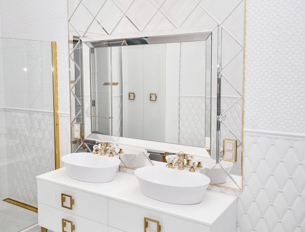 Lifestyle image of a white and gold bathroom, with a painted white double washbasin unit with gold feet and handles, gold taps and a gold bracket on the glass shower screen